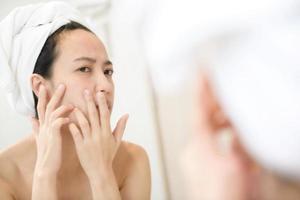 piel problemática. Mujeres asiáticas jóvenes preocupadas haciendo estallar granos en la mejilla mientras están de pie cerca del espejo en el baño. jóvenes asiáticas con acné foto