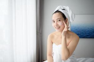 Happy young Asian woman applying face lotions while wearing a towel and touching her face. Daily makeup and skincare photo