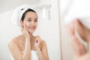 Happy young Asian woman applying face lotions while wearing a towel and touching her face in bathroom photo