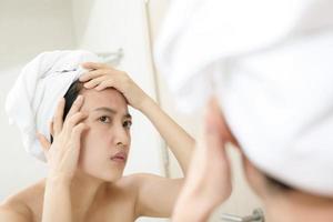 piel problemática. Mujeres asiáticas jóvenes preocupadas haciendo estallar granos en la mejilla mientras están de pie cerca del espejo en el baño. jóvenes asiáticas con acné foto