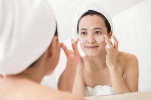 feliz joven asiática aplicando lociones faciales mientras usa una toalla y toca su cara en el baño foto