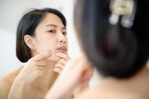 Problem skin. Concerned young asian women popping pimple on cheek while standing near mirror in bathroom. young asian women with acne photo