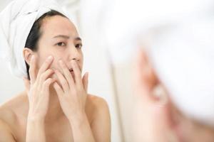 Problem skin. Concerned young asian women popping pimple on cheek while standing near mirror in bathroom. young asian women with acne photo