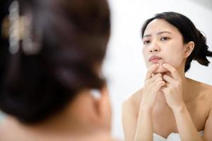 Problem skin. Concerned young asian women popping pimple on cheek while standing near mirror in bathroom. young asian women with acne photo