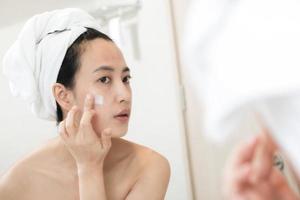 Happy young Asian woman applying face lotions while wearing a towel and touching her face in bathroom photo