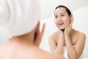 feliz joven asiática aplicando lociones faciales mientras usa una toalla y toca su cara en el baño foto