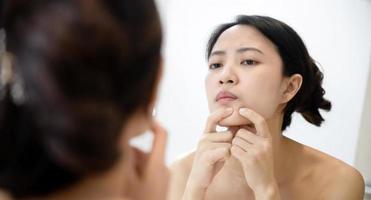 Problem skin. Concerned young asian women popping pimple on cheek while standing near mirror in bathroom. young asian women with acne photo