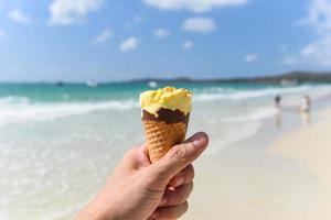 Helado derretido en la playa en verano, clima cálido, paisaje oceánico, naturaleza, vacaciones al aire libre, mango de helado amarillo con cono de helado de nueces en la mano con el mar foto
