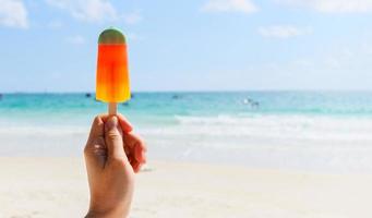 palito de helado en la mano con fondo marino - fruta de helado de colores en la playa en verano clima cálido océano paisaje naturaleza vacaciones al aire libre foto