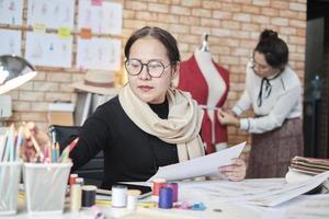 Asian middle-aged female fashion designer and young trainee tailor work in a studio with colorful thread and sewing fabric for a dress design collection ideas, professional boutique small business. photo