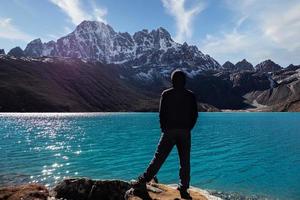 A silhouette of a man in Nepal is overlooking the Gokyo village's turquoise blue lake into the blue and cloudy sky with a big snow-capped mountain in between. photo