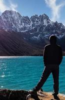 A silhouette of a man in Nepal is overlooking the Gokyo village's turquoise blue lake into the blue and cloudy sky with a big snow-capped mountain in between. photo