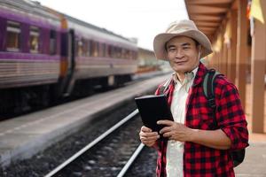 Asian man traveller is at railway station, wears hat, holds smart tablet. Concept, travel by train  in Thailand can book or by ticket online. Technology and transportation. photo