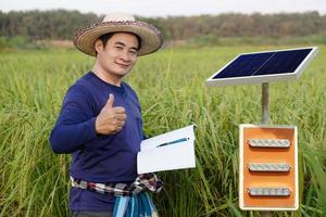 Asian man farmer wears hat, blue shirt, holds notebook paper, thumbs up, stand beside mini solar panel at paddy field. Concept, Renewable energy, Natural power in agriculture. Eco-friendly lifestyle. photo