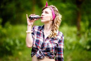beautiful young girl in a plaid shirt and short denim shorts in pin-up style in forest drinks photo