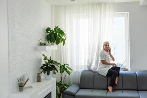 Happy beautiful business woman working on a laptop sitting near window in living room of modern home. Stay home and self isolation quarantine covid-19 concept. photo