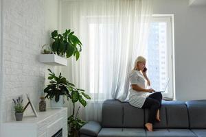 feliz hermosa mujer de negocios trabajando en una laptop sentada cerca de la ventana en la sala de estar de la casa moderna. quédese en casa y autoaislamiento concepto de cuarentena covid-19. foto