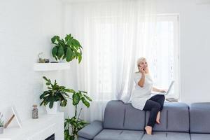 feliz hermosa mujer de negocios trabajando en una laptop sentada cerca de la ventana en la sala de estar de la casa moderna. quédese en casa y autoaislamiento concepto de cuarentena covid-19. foto