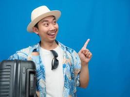 Asian man traveler hold luggage gentle smile gesture point finger blue background photo