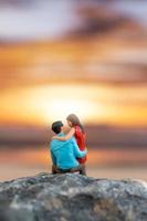 gente en miniatura, pareja sentada en una playa de mar con fondo de puesta de sol foto