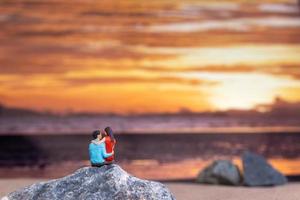 gente en miniatura, pareja sentada en una playa de mar con fondo de puesta de sol foto