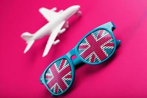 Turquoise sunglasses with United Kingdom flag in lenses on crazy pink background with white airplane. T photo