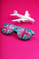 Turquoise sunglasses with United Kingdom flag in lenses on crazy pink background with white airplane. T photo