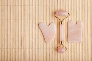 A set of tools for face Massage technique Gua Sha made of natural rose quartz. Roller, jade stone and oil in a glass jar, on a straw background for face and body care. photo