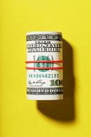 A roll of hundred-dollar American bills is tied with a red elastic band on a yellow background. photo