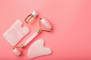 un conjunto de herramientas para la técnica de masaje facial gua sha hechas de cuarzo rosa natural sobre un fondo rosa. rodillo, piedra de jade y aceite en tarro de cristal para el cuidado facial y corporal.