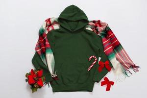 Close up green blank template hoodie copy space. Christmas Holiday concept. Top view mockup hoodie, scarf, hat. Red holidays decorations white background. Happy New Year accessories. Selective focus photo