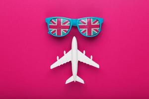 Turquoise sunglasses with United Kingdom flag in lenses on crazy pink background with white airplane. T photo