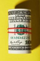 A roll of hundred-dollar American bills is tied with a red elastic band on a yellow background. photo