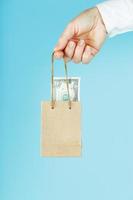 A small paper bag for financial aid and support made of paper at arm's length with US dollars on a blue background. The concept of negligible financial support in business, photo