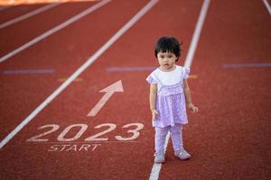 el bebé asiático comienza a correr en el nuevo año 2023. el inicio de la niña corredora corriendo en la pista de carreras de carril de entrenamiento va a la meta del éxito. personas corriendo como parte del número 2023. cuidado de la salud deportiva. foto