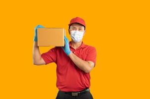 repartidor asiático con máscara quirúrgica y guantes médicos con uniforme rojo aislado en fondo amarillo, sostiene cajas de paquetes para enviar o transportar paquetes por correo. foto