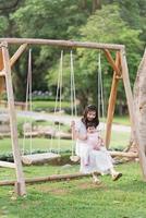 madre e hija asiáticas sentadas en un columpio y sonriendo con alegría y felicidad, buenos momentos familiares cálidos y felices. ir de vacaciones en el parque jardín o resort en natural. concepto de actividad familiar. foto