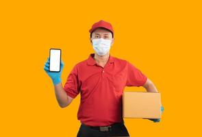 Asian delivery man worker showing mobile phone on white screen wearing surgical mask, medical gloves in red uniform isolated on yellow background, using mobile checking stock and hold parcel boxes. photo