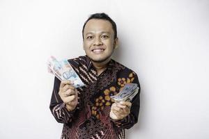 A happy young Asian man is wearing batik shirt and holding cash money in Indonesian rupiah isolated by white background photo