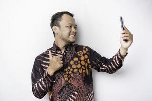un retrato de un hombre asiático feliz con un pantalones batik y sosteniendo su teléfono, aislado de fondo blanco foto
