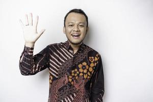 Young cheerful Indonesian man wearing batik shirt with happy positive smile waving hand hello isolated over white color background photo