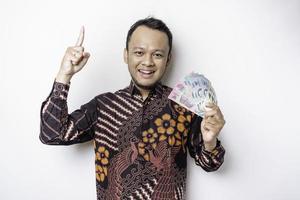 A happy young Asian man is wearing batik shirt, pointing at the copy space on top of him while holding money in Indonesian rupiah isolated by white background photo