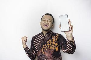 A happy young Asian man wearing batik shirt showing successful expression showing copy space on his phone isolated by white background photo