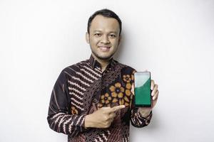 A portrait of a smiling Asian man wearing a batik shirt and showing green screen on her phone, isolated by white background photo