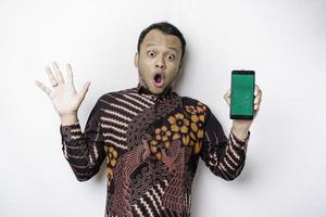 hombre asiático conmocionado con pantalones batik y mostrando una pantalla verde en su teléfono, aislado de fondo blanco foto