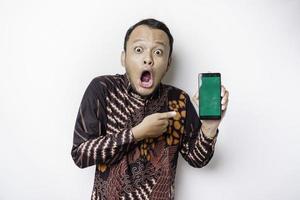 Shocked Asian man wearing batik shirt and showing green screen on his phone, isolated by white background photo