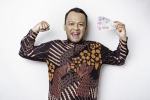 An excited young Asian man is wearing batik shirt and holding cash money in Indonesian rupiah gesturing strong pose by lifting his arms, isolated by white background photo