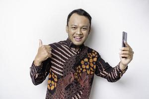 A portrait of a happy Asian man wearing batik shirt and holding his phone, isolated by white background photo