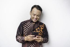 un retrato de un hombre asiático feliz con un pantalones batik y sosteniendo su teléfono, aislado de fondo blanco foto