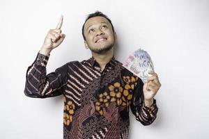 un joven asiático feliz lleva un pantalones de batik, señalando el espacio de la copia encima de él mientras sostiene dinero en rupias indonesias aislado de fondo blanco foto